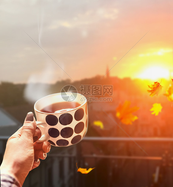 女人手着杯子,秋天的城市背景下,用热饮料蒸汽,带着飞扬的树叶夕阳的光芒图片