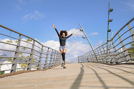 黑色女人,AFRO发型,穿着溜冰鞋,户外骑着城市桥,张开双臂阳光明媚的日子里,微笑的年轻女溜冰天空丽的云彩阿夫图片
