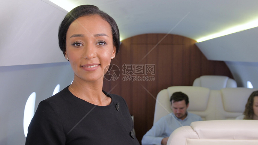 乘坐私人飞机的空姐肖像商务飞机机舱内微笑的女飞机乘务员,背景上有乘客商务飞机旅行图片