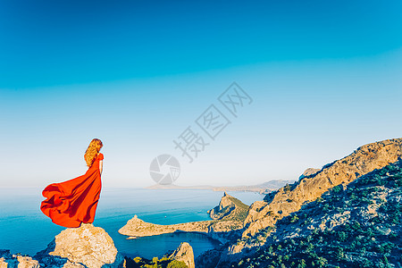 穿着红色连衣裙的年轻美女望着山海蓝天背景上的自然女孩时尚照片图片