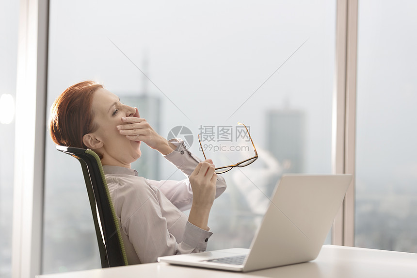 疲惫的年轻女商人打着哈欠,坐办公室的桌子上着笔记本电脑图片