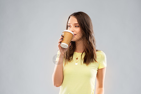 外卖人们的轻的女人十几岁的女孩穿着T恤,喝着灰色背景的咖啡轻女子十几岁的女孩喝咖啡图片
