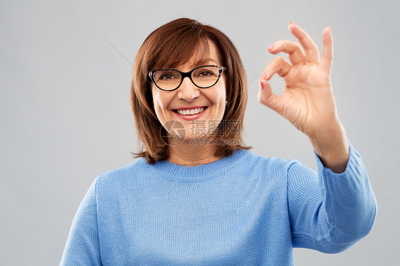 手势,视觉老人的微笑的老年女子眼镜上的肖像,灰色背景上OK手标志快乐的老年女子戴着眼镜,出好的手势图片