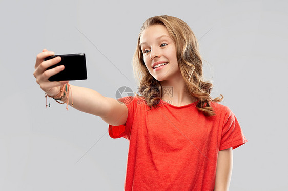 技术人的微笑的轻妇女十几岁的女孩穿着空白的红色T恤,灰色背景下智能手机自拍微笑的十几岁女孩用智能手机自拍图片