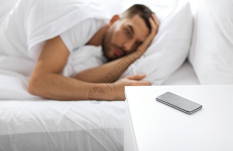 技术人的智能手机床头柜附近的轻人睡家里的床上床头柜上的智能手机靠近熟睡的男人图片