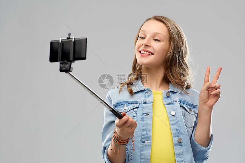 技术人的快乐的微笑少女用智能手机自拍杆上拍照,并灰色背景上表现出平十几岁的女孩用智能手机自拍图片