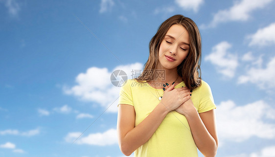 欣赏,情人节真诚的情感感谢轻的女人十几岁的女孩,闭着眼睛,穿着t恤,双手抱着胸部心脏,蓝天云彩的背景下图片