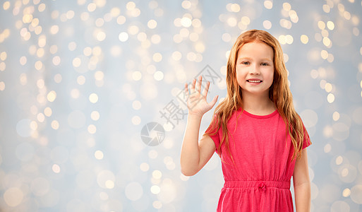 童人们的微笑的红头发女孩穿着粉红色的裙子,挥舞着手节日的灯光背景微笑的红头发女孩挥手关灯图片