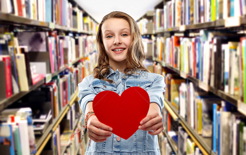 情人节,爱教育理念微笑的漂亮少女穿着牛仔夹克,图书馆背景上捧着红色的心微笑的十几岁女孩,红色的心图书馆上空图片