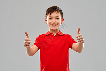 童,手势人的微笑的小男孩红色马球T恤拇指灰色背景穿着红色T恤的微笑男孩竖大拇指背景图片