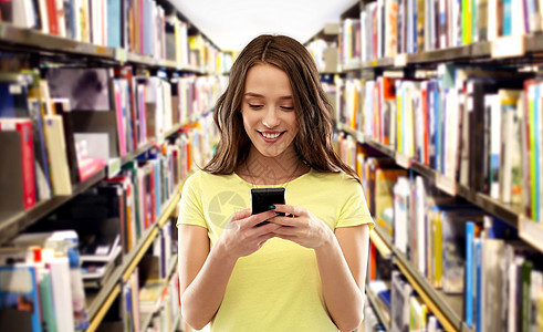 技术教育人的微笑的轻妇女十几岁的女孩穿着空白的黄色T恤,图书馆背景的书架上用智能手机十几岁的学生女孩图书图片