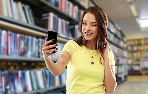技术人的微笑的轻妇女十几岁的女孩穿着空白的黄色T恤,图书馆背景下智能手机自拍微笑的十几岁女孩用智能手机自拍图片