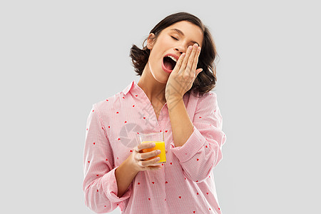 人们早晨的快乐的昏昏欲睡的打哈欠的轻妇女睡衣与杯橙汁灰色的背景快乐的哈欠轻的女人睡衣与果汁图片
