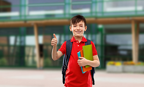 红色大拇指教育人的微笑的小学生男孩穿着红色马球T恤,戴着眼镜,带着书书包,学校院子的背景上竖大拇指学生男孩着书书包竖背景