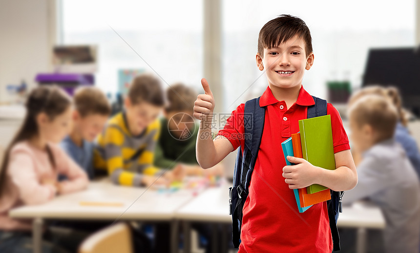 教育人的微笑的小学生男孩穿着红色马球T恤,戴着眼镜,带着书书包,学校的课堂背景上竖大拇指学生男孩着书书包竖图片