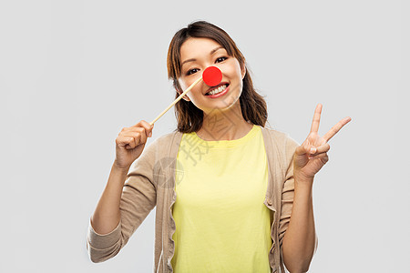 红鼻子日,派道具照片展位快乐的亚洲轻妇女与小丑鼻子平灰色背景红色小丑鼻子的亚洲女人表现出平图片