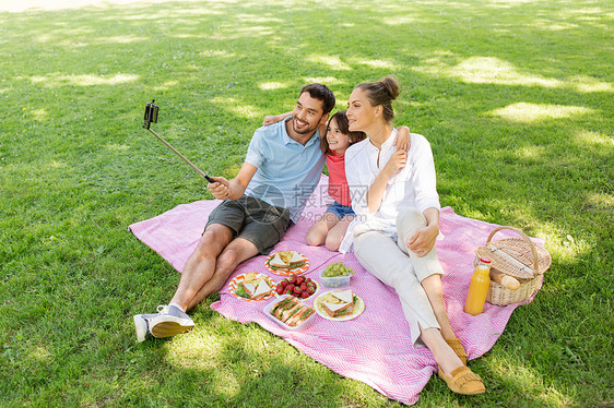 家庭休闲技术快乐的母亲父亲女儿夏季公园用智能手机自拍杆上野餐拍照家人公园野餐自拍图片