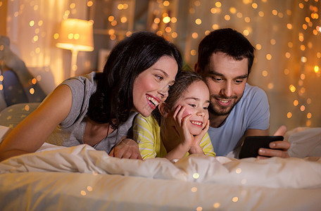 准爸爸人们家庭的快乐的母亲,父亲小女儿,晚上家里睡觉时带着智能手机快乐的家庭,晚上床上智能手机背景