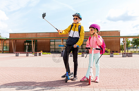 教育,童人的快乐的学校孩子戴着头盔,背包骑着滑板车,户外自拍杆上用智能手机拍照快乐的学校孩子与滑板车自拍图片