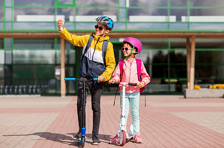 教育,童人的快乐的学校孩子戴着头盔,背包骑着滑板车,户外用智能手机自拍快乐的学校孩子与滑板车自拍图片