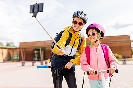 教育,童人的快乐的学校孩子戴着头盔,背包骑着滑板车,户外自拍杆上用智能手机拍照快乐的学校孩子与滑板车自拍图片