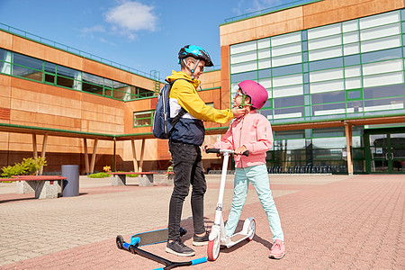 安全,童护理快乐的学校男孩与背包紧固女孩的头盔滑板车骑学校男孩为滑板车系紧女孩的头盔背景图片