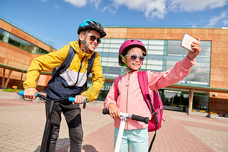 教育,童人的快乐的学校孩子戴着头盔,背包滑板车,户外用智能手机自拍快乐的学校孩子与滑板车自拍图片