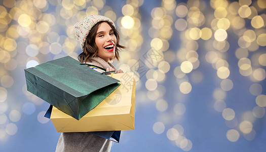 诞节,季节销售消费主义快乐的微笑轻妇女针冬帽毛衣与购物袋节日灯上的蓝色背景诞节戴着购物袋的戴帽子的女人背景图片