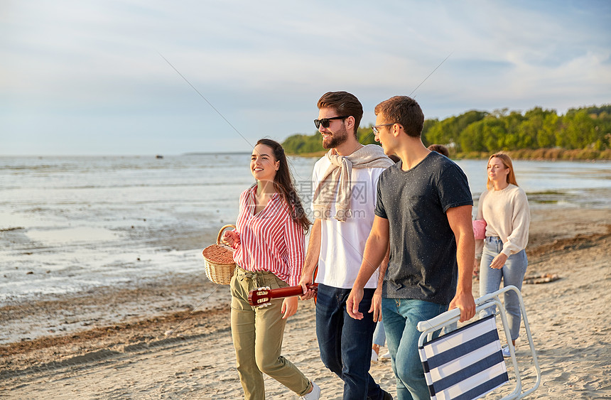 友谊休闲人的群快乐的朋友带着吉他野餐篮折叠椅夏天沿着海滩散步快乐的朋友沿着夏天的海滩散步图片