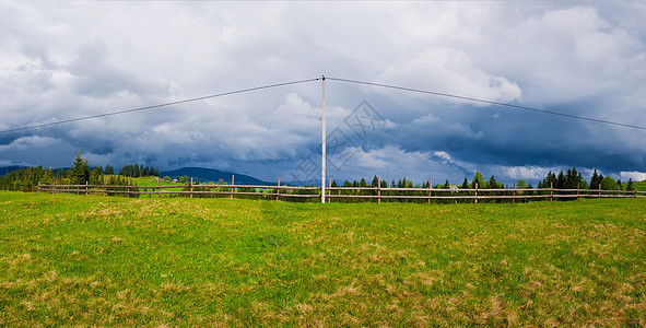 电力传输杆山上,电缆沿木轨围栏通向村庄全景景观,绿色的田野多云的天空背景图片