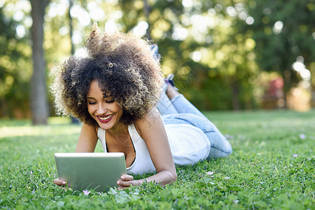 轻的黑人妇女,留着AFRO的发型,躺城市公园的草地上,看着她的平板电脑穿着休闲衣服的混合女孩图片