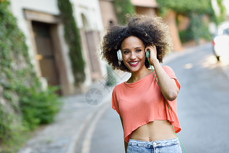 城市背景中轻迷人的黑人女孩的肖像,用耳机听音乐女人穿着皮夹克蓝色牛仔裤,留着AFRO的发型图片