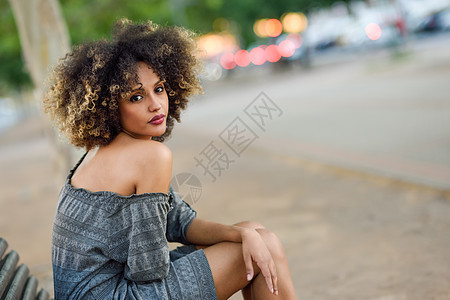 轻的黑人女孩,留着AFRO发型,坐城市背景的长凳上穿着休闲衣服的混合女人图片