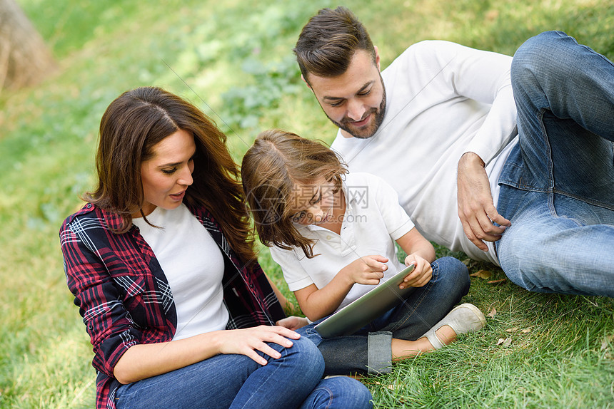 城市公园玩平板电脑的幸福家庭爸爸妈妈小女儿坐草地上笑图片