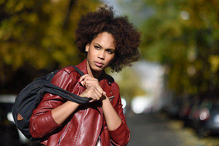 轻的黑人女,留着AFRO发型,站条城市街道上,背着个包混合女人穿红色皮夹克白色连衣裙与城市背景图片