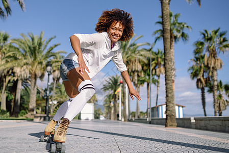 微笑的黑人妇女溜冰鞋骑户外海滩长廊与棕榈树阳光明媚的日子里,微笑的女孩留着AFRO的发型图片