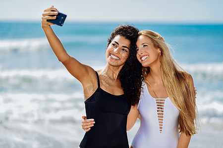 两个女人海滩上用智能手机自拍两位轻女热带海滩上穿着泳衣用智能手机自拍图片