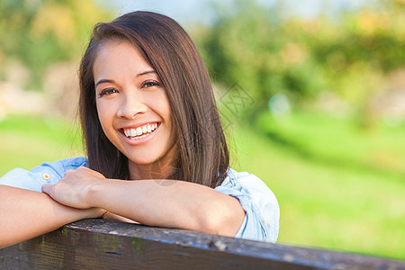 美丽快乐的亚洲欧亚青妇女女孩穿着牛仔衬衫,微笑与完美的牙齿,阳光下靠篱笆上图片