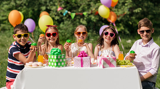 假期,童庆祝快乐的孩子戴着太阳镜坐桌子上的生日聚会夏季花园吃纸杯蛋糕孩子们夏天的生日聚会上吃纸杯蛋糕孩图片