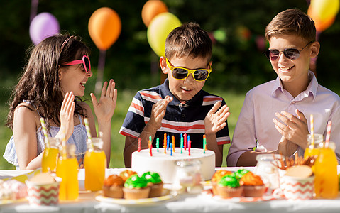 生日party假期,童庆祝快乐的孩子蛋糕上着蜡烛,坐夏天的花园聚会上,为生日男孩鼓掌快乐的孩子夏天的生日聚会上吃蛋糕背景