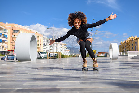 轻合身的黑人妇女溜冰鞋骑户外的城市街道上张开双臂阳光明媚的日子里,微笑的女孩留着AFRO的发型图片