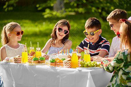 假期,童庆祝快乐的孩子与蜡烛生日蛋糕上坐桌子上的夏季花园聚会快乐的孩子夏天的生日聚会上吃蛋糕图片