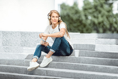 快乐的轻女,带着老式音乐耳机带走的咖啡杯,平板电脑上上网,听音乐,坐城市背景下的楼梯上图片