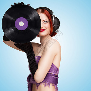 张美丽感女孩的彩色照片,她戴着老式音乐耳机,手里着蓝色背景的紫色LP微槽乙烯基唱片图片