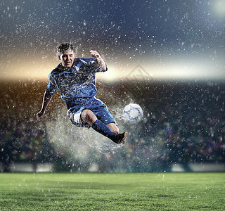 足球运动员击球穿着蓝色衬衫的足球运动员雨下体育场把球打得高高的图片