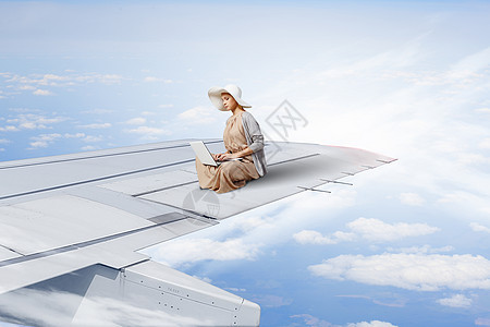 绝穿着连衣裙帽子的女人坐飞机机翼上,笔记本电脑上工作图片