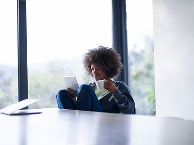 轻的非裔美国妇女微笑着坐明亮的窗户旁边,看着平板电脑,着白色的杯子她豪华的家图片