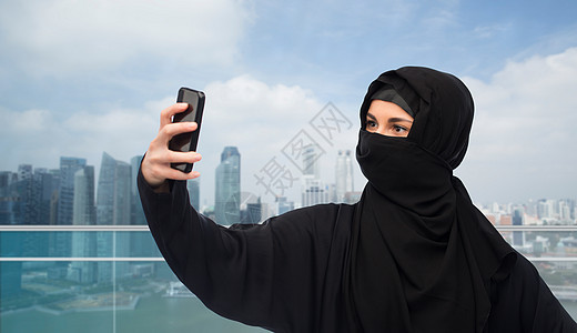 技术人的穆斯林妇女头巾自拍与智能手机新加坡城市摩天大楼背景穆斯林妇女戴着头巾,用智能手机自拍图片