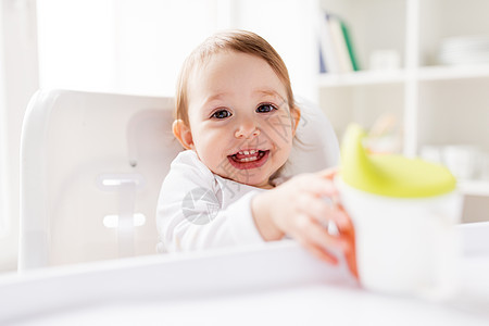 童人们的快乐的微笑小婴儿喝喷口杯坐高椅子家里婴儿家里的高椅子上喷口杯里喝水图片