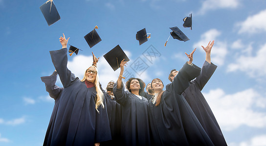 教育,毕业人的群快乐的国际学生穿着学士学位礼服,蓝天云彩的背景上投掷迫击炮板快乐的学生把沙浆板扔上来图片